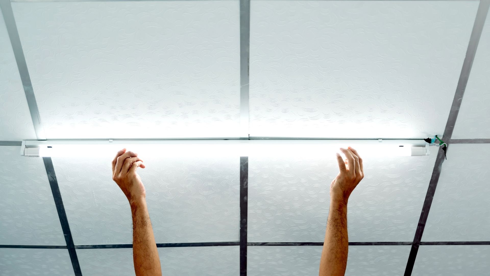 Two arms reaching up towards a bright, grid-like ceiling light
